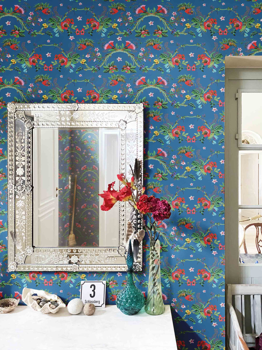 A wall decorated with a blue floral chinoiserie wallpaper, featuring a mirror and a vase with flowers in the foreground.