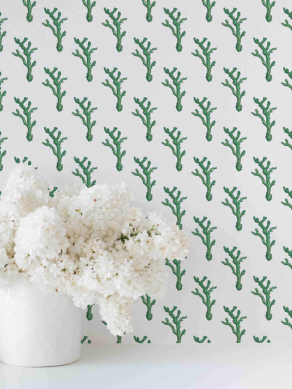 A wall with wallpaper with a green coral dash pattern, next to a white vase with white flowers.