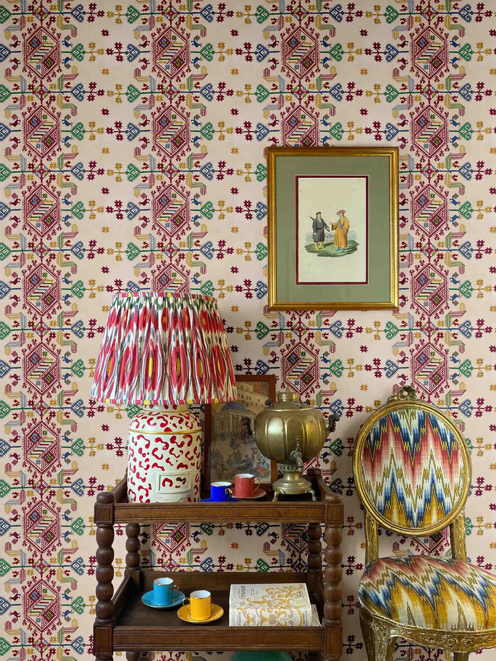 A wall decorated with oat cream colored wallpaper featuring a geometric pattern, in a room setting with a chair covered in a colorful geometric pattern, a lamp, and a small table with accessories.
