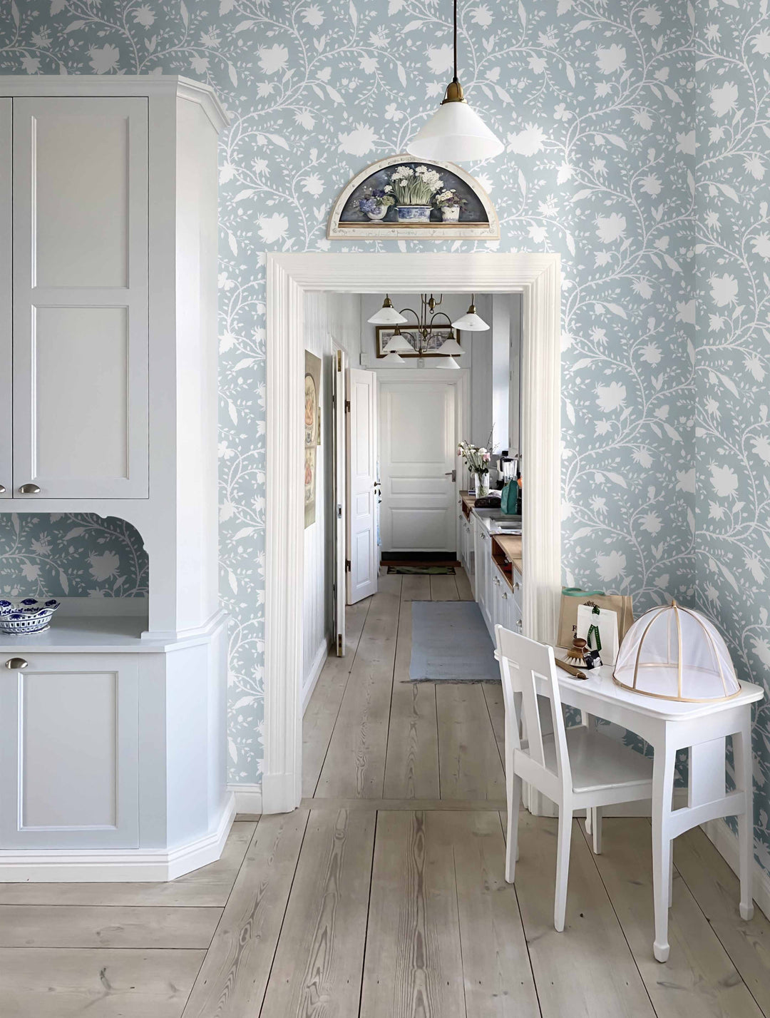 A room with light blue floral wallpaper and a white wood floor, featuring a kitchen setting, and a table in the foreground.