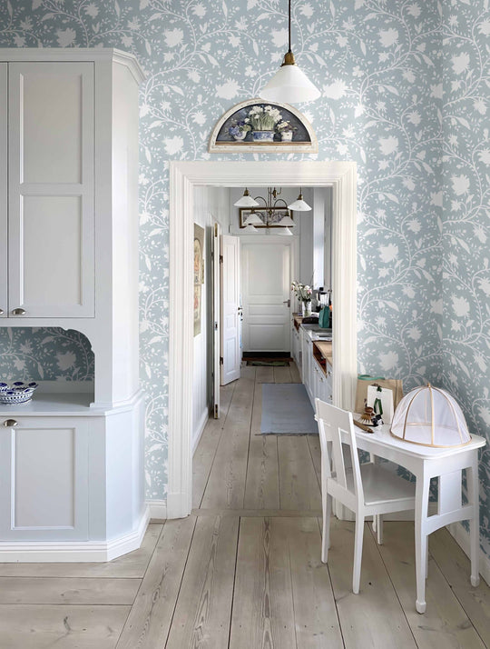 A room with light blue floral wallpaper and a white wood floor, featuring a kitchen setting, and a table in the foreground.