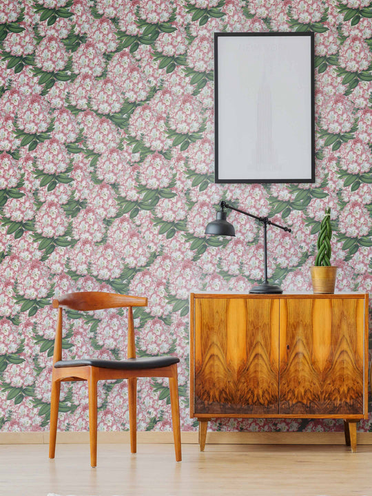 A room with  bold floral wallpaper featuring pink flowers, a chair, a mirror, and a wooden cabinet with a lamp.